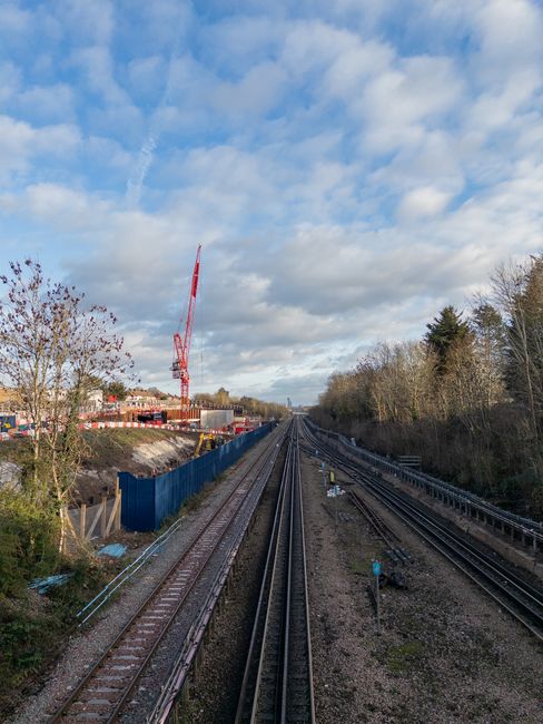 A panoramic view of parallel railway tracks extending into the distance beneath a partly cloudy sky, with construction activity visible on the left side where a tall red crane and blue safety barriers