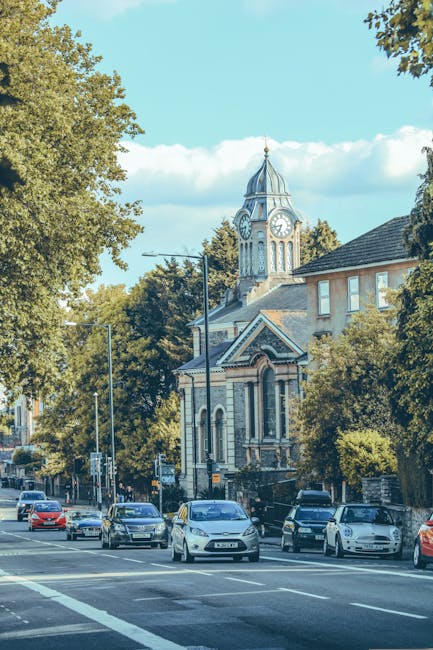 A street scene in Ealing with multiple parked cars along the curb, including sedans and hatchbacks, in front of a historic church building with a prominent clock tower, which features a pointed spire and arched windows. Tall trees with dense green foliage line both sides of the road, casting shadows on the pavement, and the sky is clear with a few clouds. Near the background, a crane is visible behind the church, indicating ongoing construction or renovation. The scene is well-lit, with ambient sunlight highlighting the details of the buildings and street environment. This image relates to house removals and moving services offered by Removal Van Ealing, illustrating the local area for logistics planning regarding home relocation and furniture transport through narrow streets typical of Ealing's urban setting.