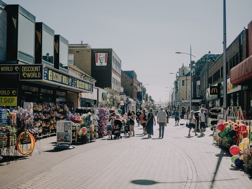 A busy pedestrian street in Ealing with various retail stores and kiosks on either side. The scene shows people walking, shopping, and pushing strollers, with some wearing casual summer clothing. On the left side, there are outdoor stalls displaying an array of colourful goods, including balloons, toys, and household items, with cardboard and plastic packaging materials visible around the displays. To the right, storefronts include retail outlets and food outlets, some with awnings or signs. The street is paved with light-coloured bricks and features lampposts lining the sidewalk. The sky is clear, indicating daylight, and the overall setting suggests an active shopping area suitable for neighbourhood relocations or home moves, where furniture transport and packing preparations might take place before loading into removal vans, such as those operated by Removal Van Ealing, in narrow street environments.