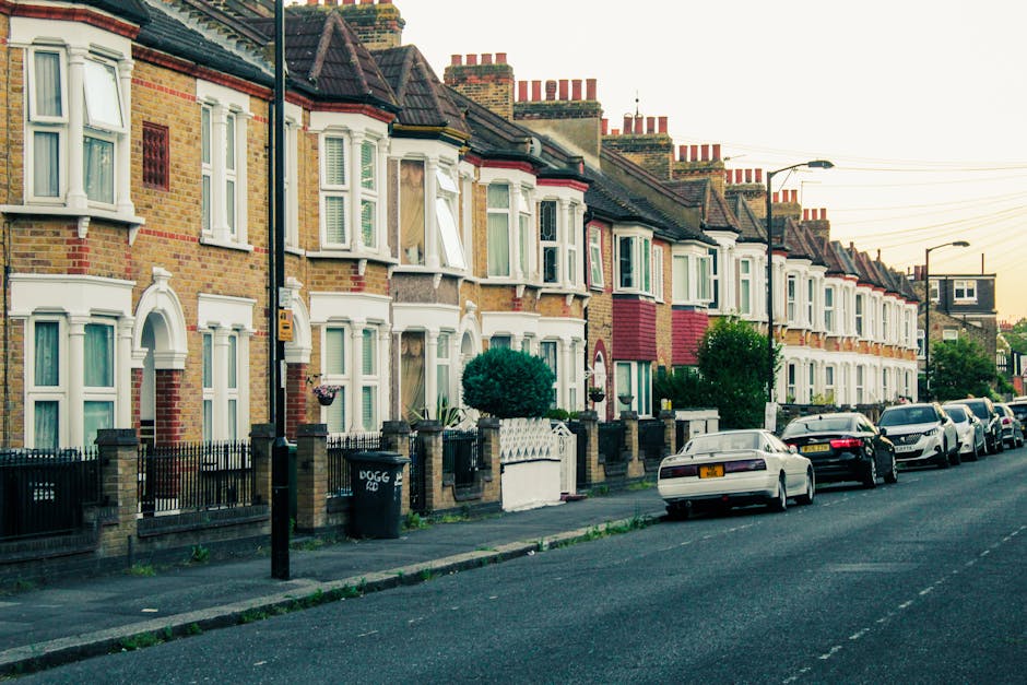 A street scene in Ealing with multiple parked cars along the curb, including sedans and hatchbacks, in front of a historic church building with a prominent clock tower, which features a pointed spire and arched windows. Tall trees with dense green foliage line both sides of the road, casting shadows on the pavement, and the sky is clear with a few clouds. Near the background, a crane is visible behind the church, indicating ongoing construction or renovation. The scene is well-lit, with ambient sunlight highlighting the details of the buildings and street environment. This image relates to house removals and moving services offered by Removal Van Ealing, illustrating the local area for logistics planning regarding home relocation and furniture transport through narrow streets typical of Ealing's urban setting.
