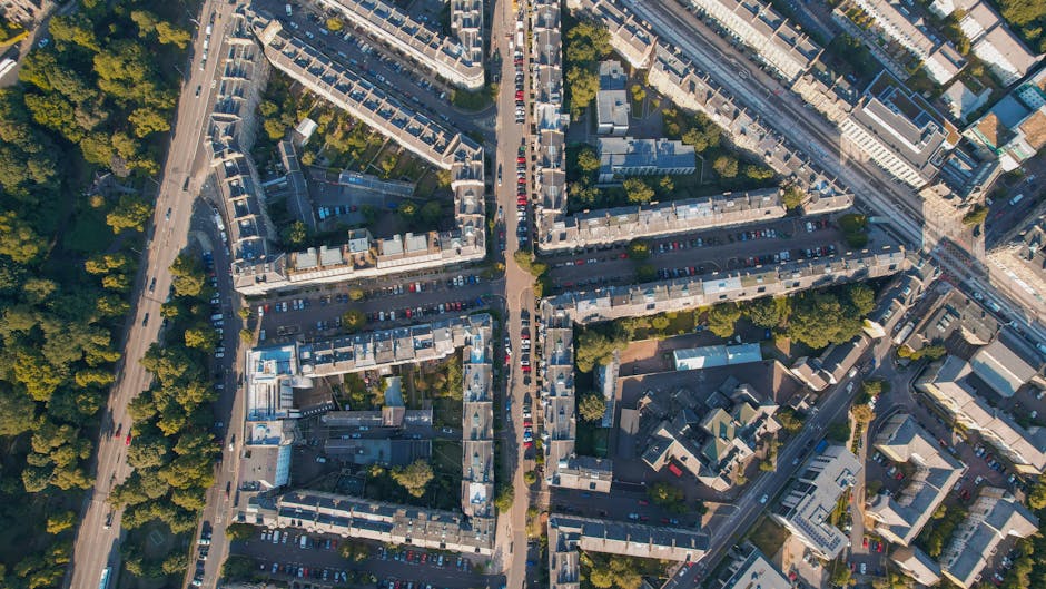 Aerial view of a residential street showing bypassing roads crossing over a railway track on a bridge, with parked cars along the curbs and houses with varying roof styles and gardens on both sides. G