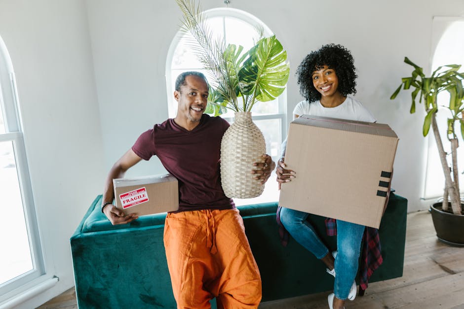 A man and woman are inside a bright, modern living room preparing for a house move, with natural light streaming through large arched windows in the background. The man, wearing a maroon t-shirt and orange trousers, holds a small cardboard box marked with a red 'Fragile' label in one hand and a large decorative vase with green monstera leaves in the other. The woman, dressed in a white top and blue jeans, carries a sizable cardboard box, possibly containing household items or packing supplies. They are seated on a dark green sofa, surrounded by other packing materials and furniture, indicating the process of home relocation. Visible in the surroundings are wooden flooring, a potted plant, and the window behind them, suggesting a typical interior setup during furniture transport or packing and moving preparations. This image reflects aspects of professional removals and house relocation services that Removal Van Ealing offers, involving packing, handling, and loading of belongings for a local move on Pitshanger Lane.