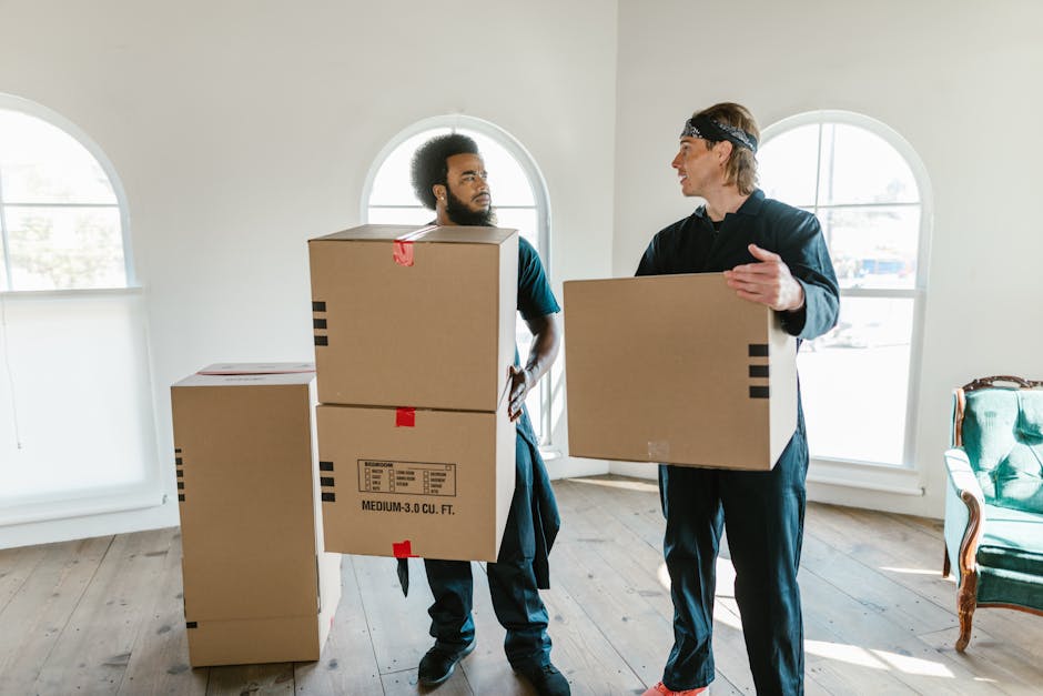 A young woman with long dark hair, wearing a white tank top and beige pants, is sitting on a wooden floor inside a residential property, surrounded by numerous large cardboard moving boxes in various